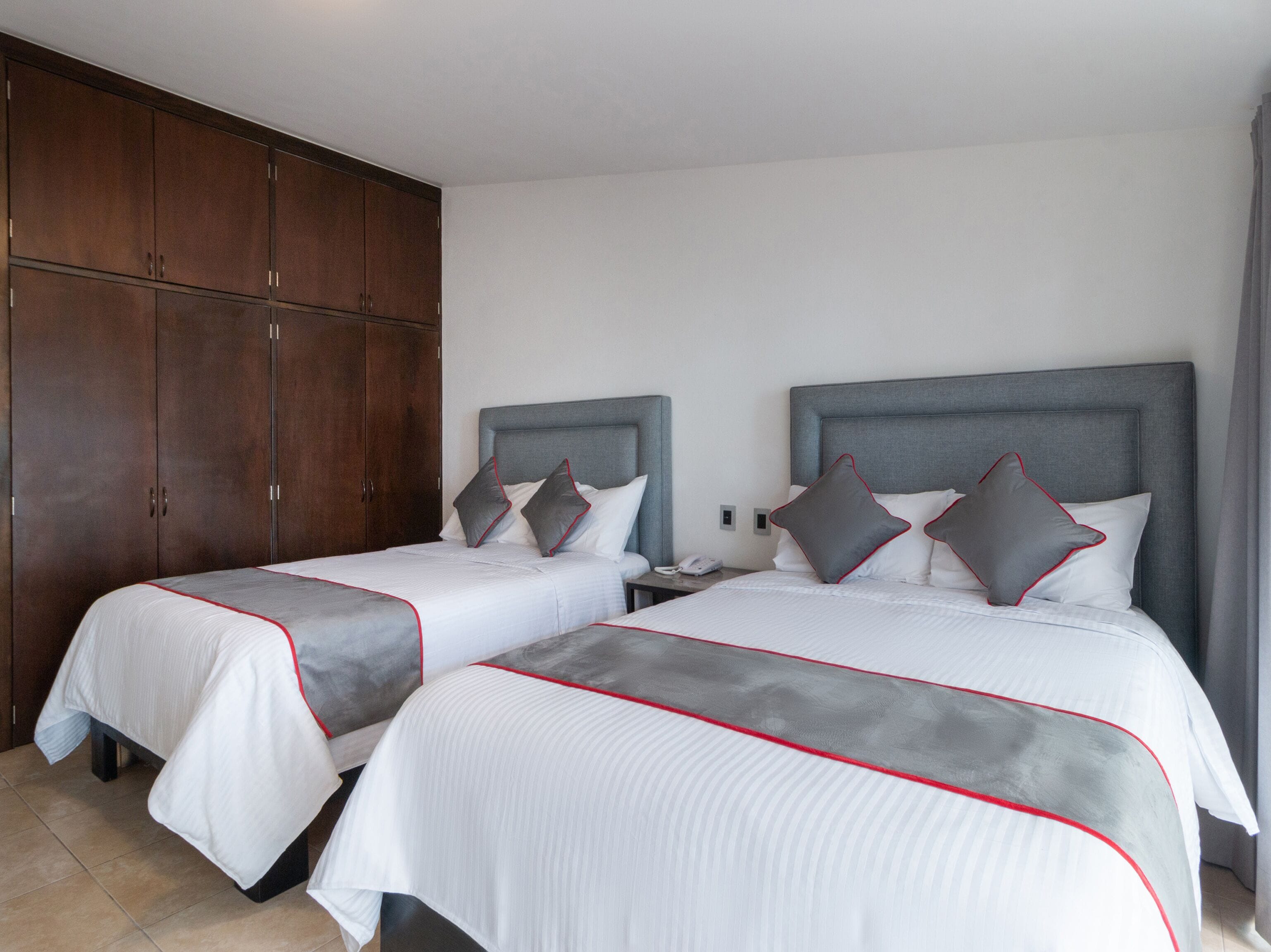 A hotel room with two beds, each with grey and white bedding and red trim. There is a wooden wardrobe and a nightstand with a phone.