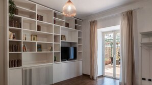 Una sala de estar con una estantería blanca, un televisor de pantalla plana, un radiador y un ventanal con cortinas.