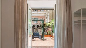Vista a través de una puerta a un patio exterior con una pérgola metálica verde, dos sillas, plantas en macetas y un suelo de baldosas.