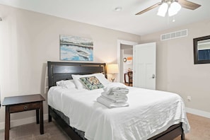 A neatly made bed with white linens and a folded towel, a small bedside table, a painting of a boat on water, a ceiling fan, and a door leading to another room.