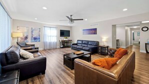 A spacious living room with a dark leather sofa, a brown leather couch, and a coffee table. There is a ceiling fan, a flat-screen TV mounted on the wall, and a painting above the sofa.