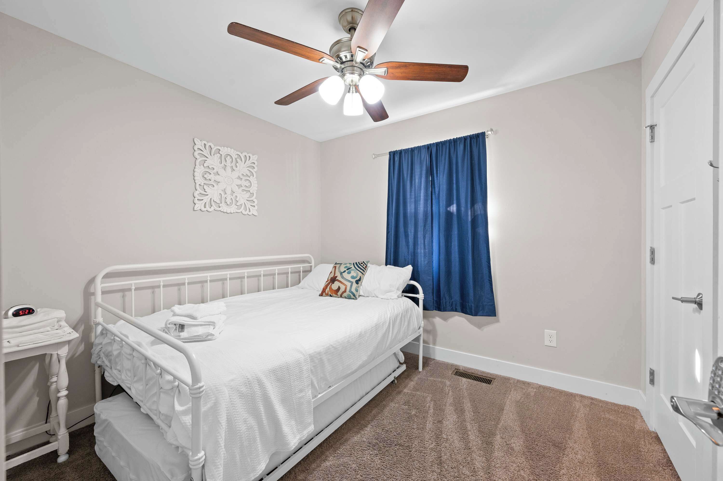 A neatly made bed with a white comforter and pillows, a ceiling fan, a wall clock, and a window with blue curtains.