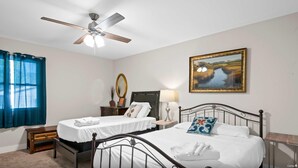 A bedroom with two single beds, a ceiling fan, a bedside table with a lamp, a dresser, a mirror, and a framed landscape painting.