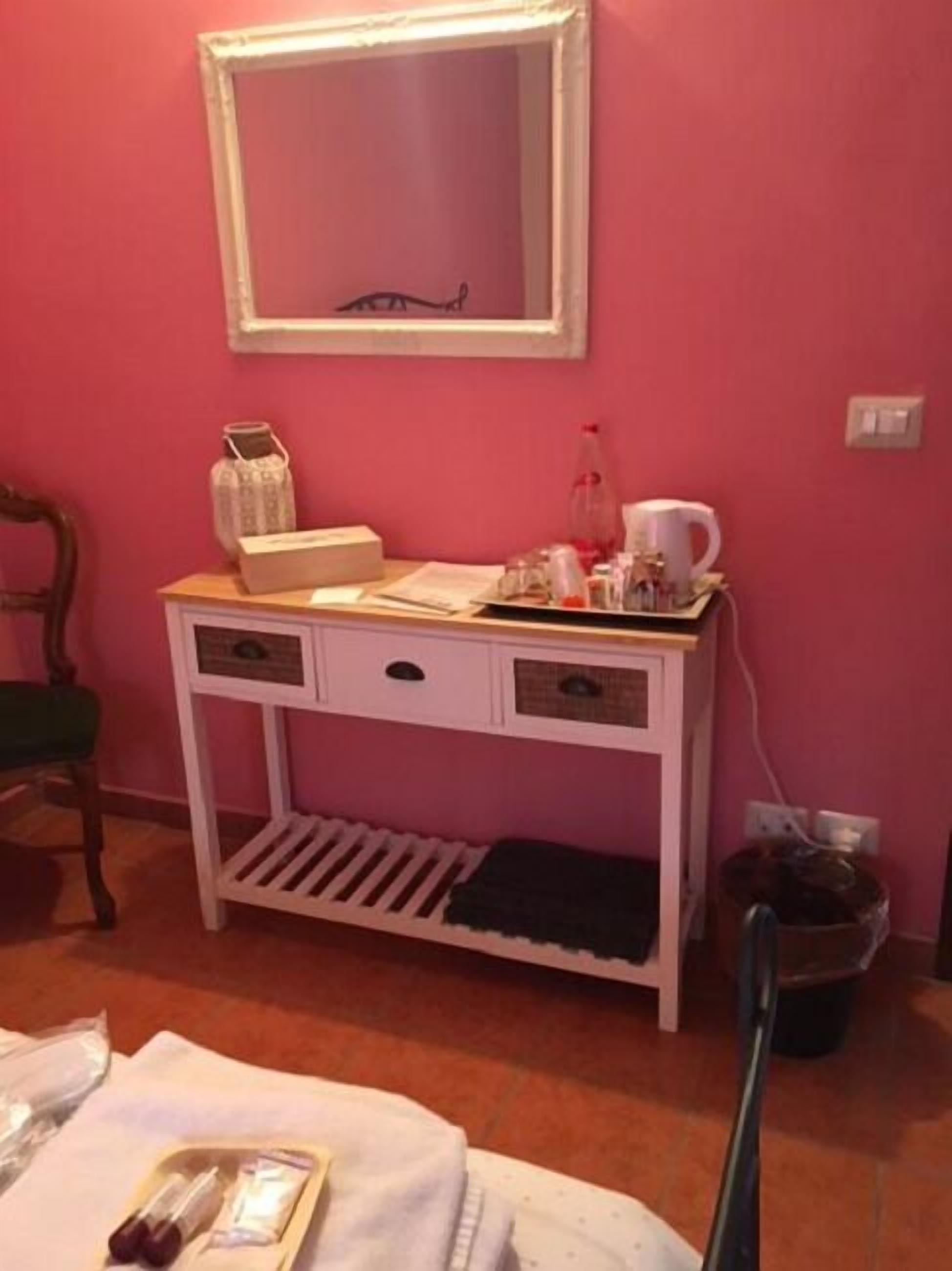 A pink-walled room with a white bedside table, a mirror, and a small shelf.