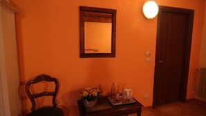 Een kamer met een houten stoel, een klein tafeltje met een mand vol bloemen, een inlijst spiegel en een deur met een lamp erboven.