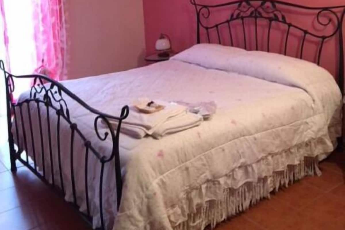 Un dormitorio con estructura de cama metálica, ropa de cama blanca, pared rosa y una ventana con cortinas rosas.