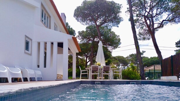 A swimming pool with a tiled edge, surrounded by white chairs and a table with umbrellas.