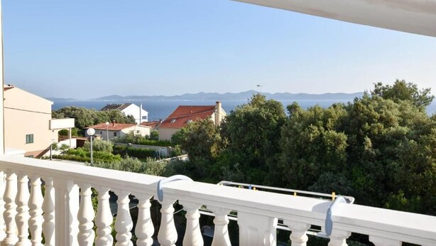 Un balcón con barandilla blanca que da a una zona residencial con casas y árboles, y una vista despejada del mar y montañas distantes.