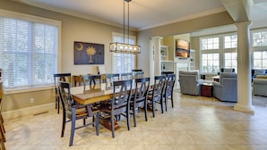 A spacious dining area with a large wooden table and chairs, a hanging light fixture, and a view into the living room.