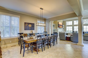 A spacious dining area with a large wooden table and chairs, a hanging light fixture, and a view into the living room.