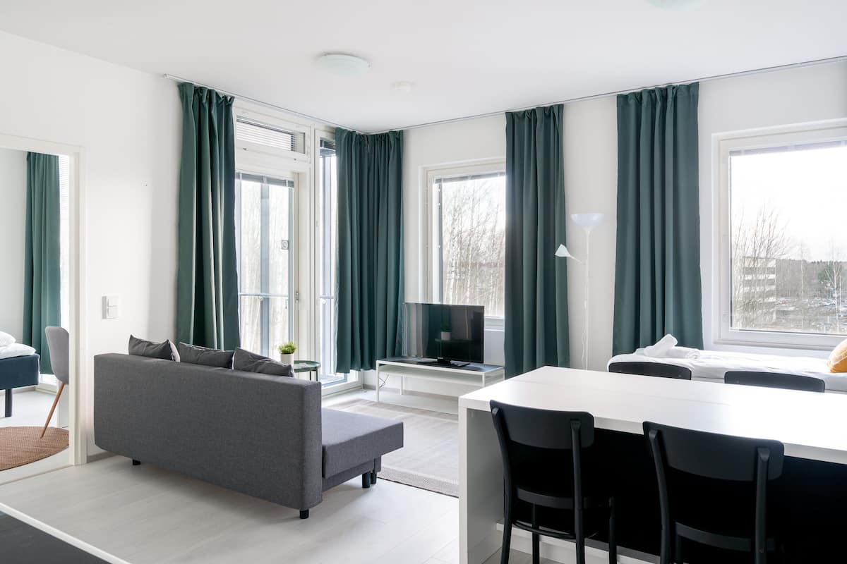 Ein modernes Wohnzimmer mit einem grauen Sofa, einem Esstisch mit Stühlen, einem Fernseher auf einem Schrank und großen Fenstern mit grünen Vorhängen.