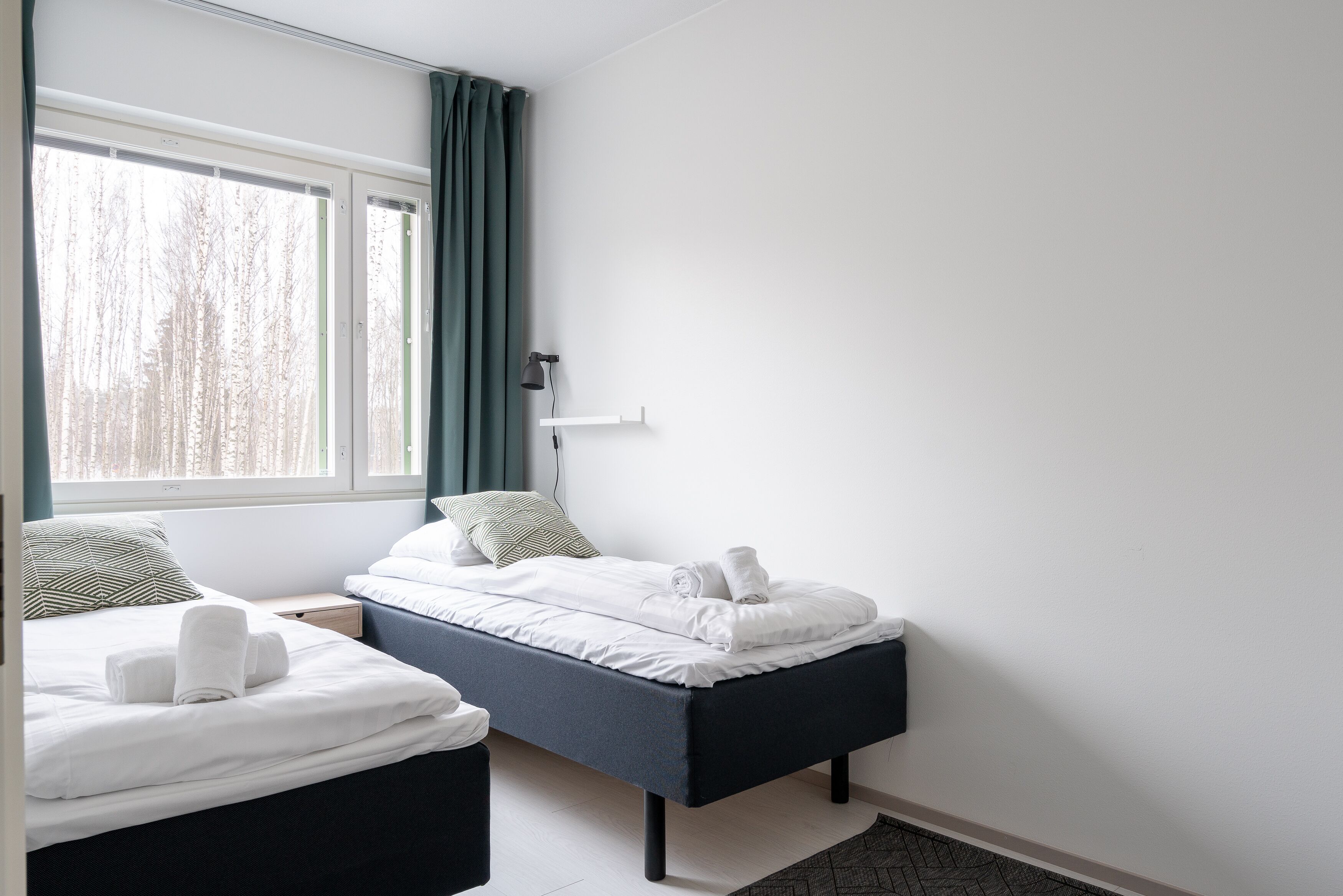 A modern hotel room with two beds, a large window with curtains, a bedside table, and a lamp.