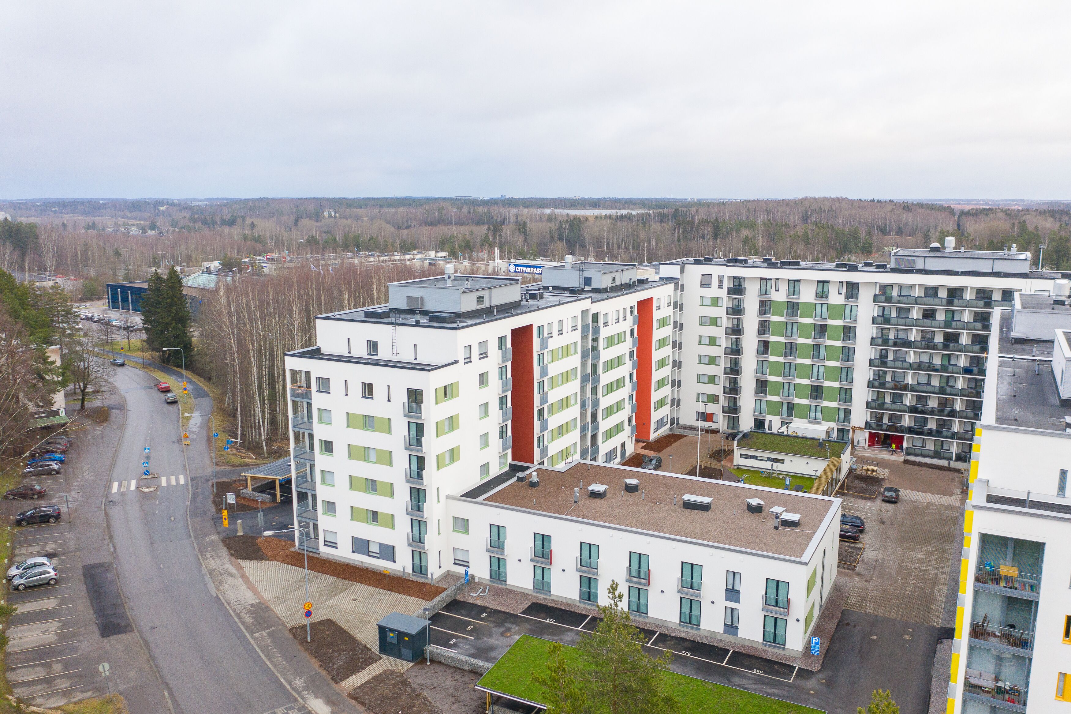 En flereetagers boligbygning med fladt tag, parkeringsområde og udsigt til et skovområde i baggrunden.