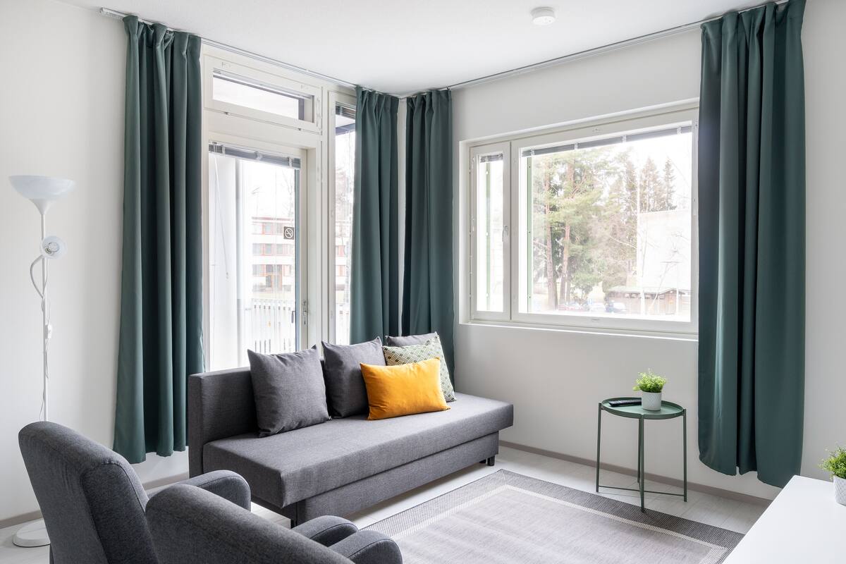 Ein modernes Wohnzimmer mit einem grauen Sofa, dunkelgrünen Vorhängen und einem kleinen runden Tisch mit einer Pflanze.