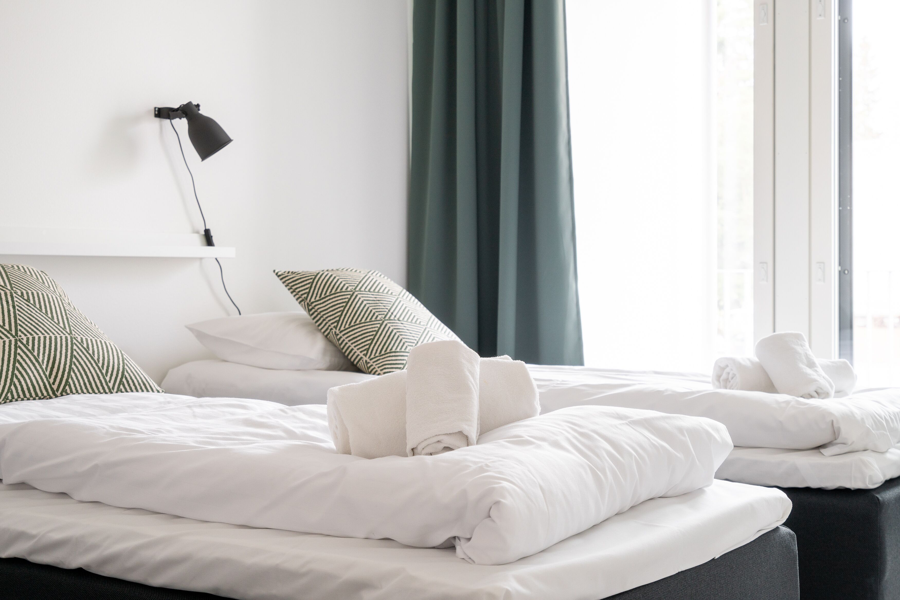A hotel room with two beds, white bedding, a black lamp on a wall-mounted shelf, and green curtains.