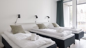 Une chambre d’hôtel avec trois lits, chacun étant équipé de draps blancs et d’une serviette. Des lampes noires sont fixées au mur au-dessus des lits.