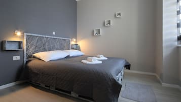 A neatly made bed with a dark quilt, two pillows, and two towels on the bed. There are wall-mounted lights and a small shelf with a plant.