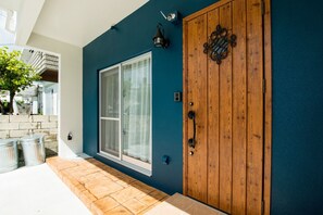 Une maison avec une porte en bois, une grande fenêtre et une petite poubelle extérieure.