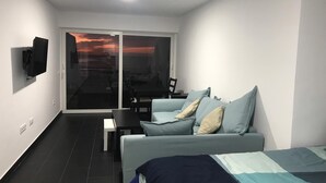 A modern bedroom with a sofa, a bed, a TV, and a view of the sunset through a glass door. - Canteras Reef - First sea line (Las Palmas de Gran Canaria)