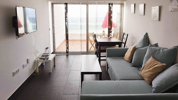 A modern living room with a sofa, a TV mounted on the wall, and a sliding glass door leading to a balcony with a table and chairs. - Canteras Reef - First sea line (Las Palmas de Gran Canaria)