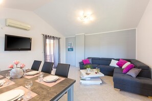 A modern living room with a dining area, a grey sofa with colorful cushions, a flat-screen TV, and a ceiling light fixture.