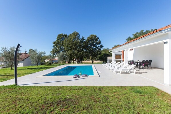 A swimming pool with lounge chairs and a barbecue area, surrounded by a grassy lawn and trees.
