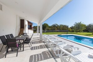 A covered outdoor area with a pool, a dining table with chairs, and a staircase leading to a house.