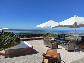 Una terrazza con mobili da esterno, tra cui una poltrona relax, un tavolo in legno e due ombrelloni bianchi che offrono ombra.