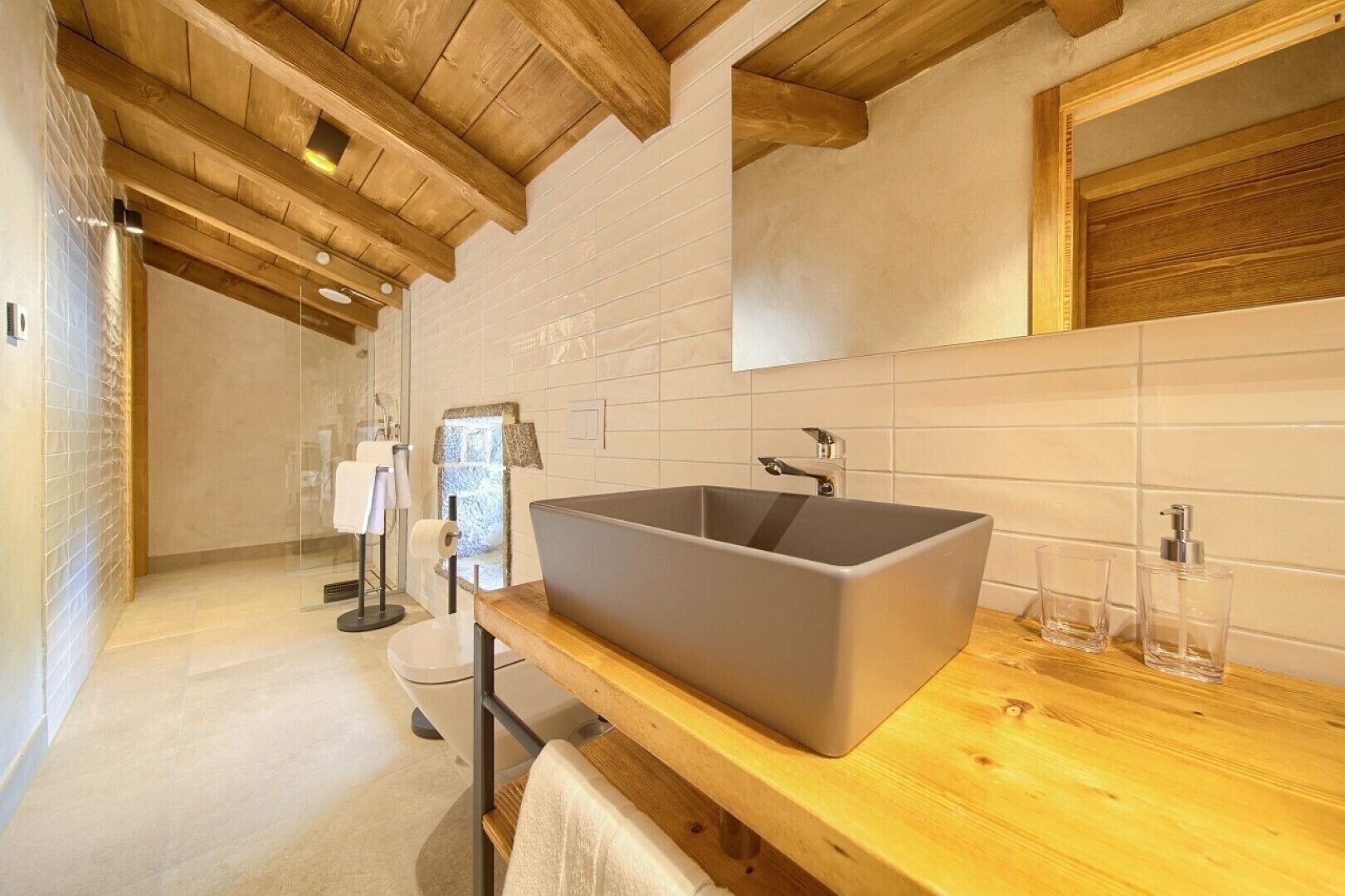 Une salle de bain moderne avec un grand lavabo rectangulaire, des poutres en bois au plafond et un mur recouvert de carreaux.