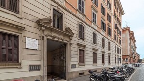 A row of parked motorcycles along a street with a building featuring classical architecture and a signboard. - GuestHost - Testaccio Art & Design Flat BOOK NOW! (Roma)