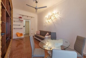 A modern living room with a glass table, grey and orange chairs, a sofa, a ceiling fan, and a wall-mounted light fixture.