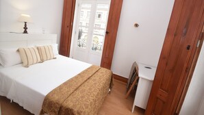 Habitación de hotel con cama, mesita de noche, lámpara, puerta de madera y vista de edificios a través de un ventanal.