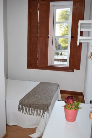 A small, neatly arranged room with a single bed, a window with wooden shutters, a white table with a pink vase, and a wooden floor.