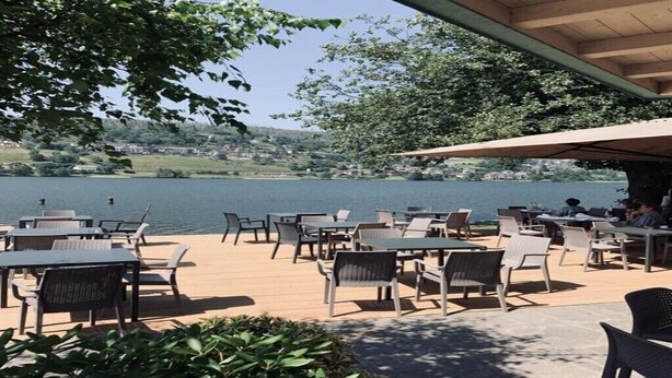 An outdoor patio with tables and chairs overlooking a lake, surrounded by greenery and a wooden deck. - Apartment "Casa Ester" 4 people (Baselga di Pinè, Italia)