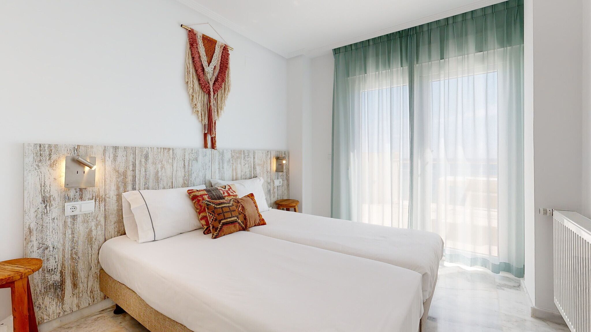 A bedroom with a large bed, a wooden headboard, two bedside lamps, and a view of the outdoors through sheer curtains.