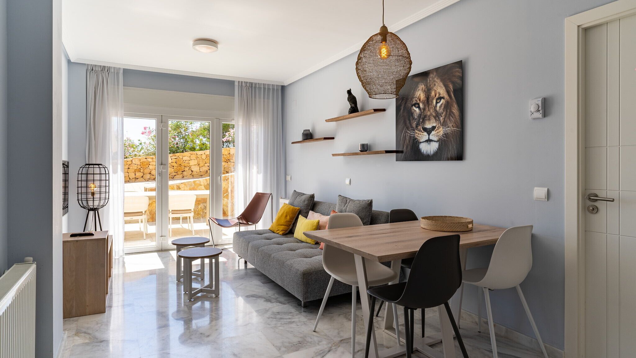 Una sala de estar moderna con un sofá gris, una mesa de comedor de madera y un ventanal con vistas a una pared de piedra y vegetación.