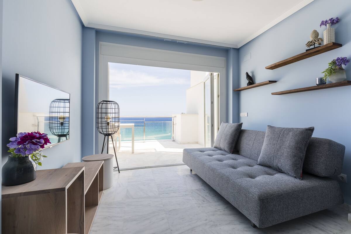 Een moderne woonkamer met een grijze bank, houten bijzettafels en een grote spiegel. Er zijn zwevende planken met decoratieve items en een uitzicht op de oceaan door grote glazen deuren.