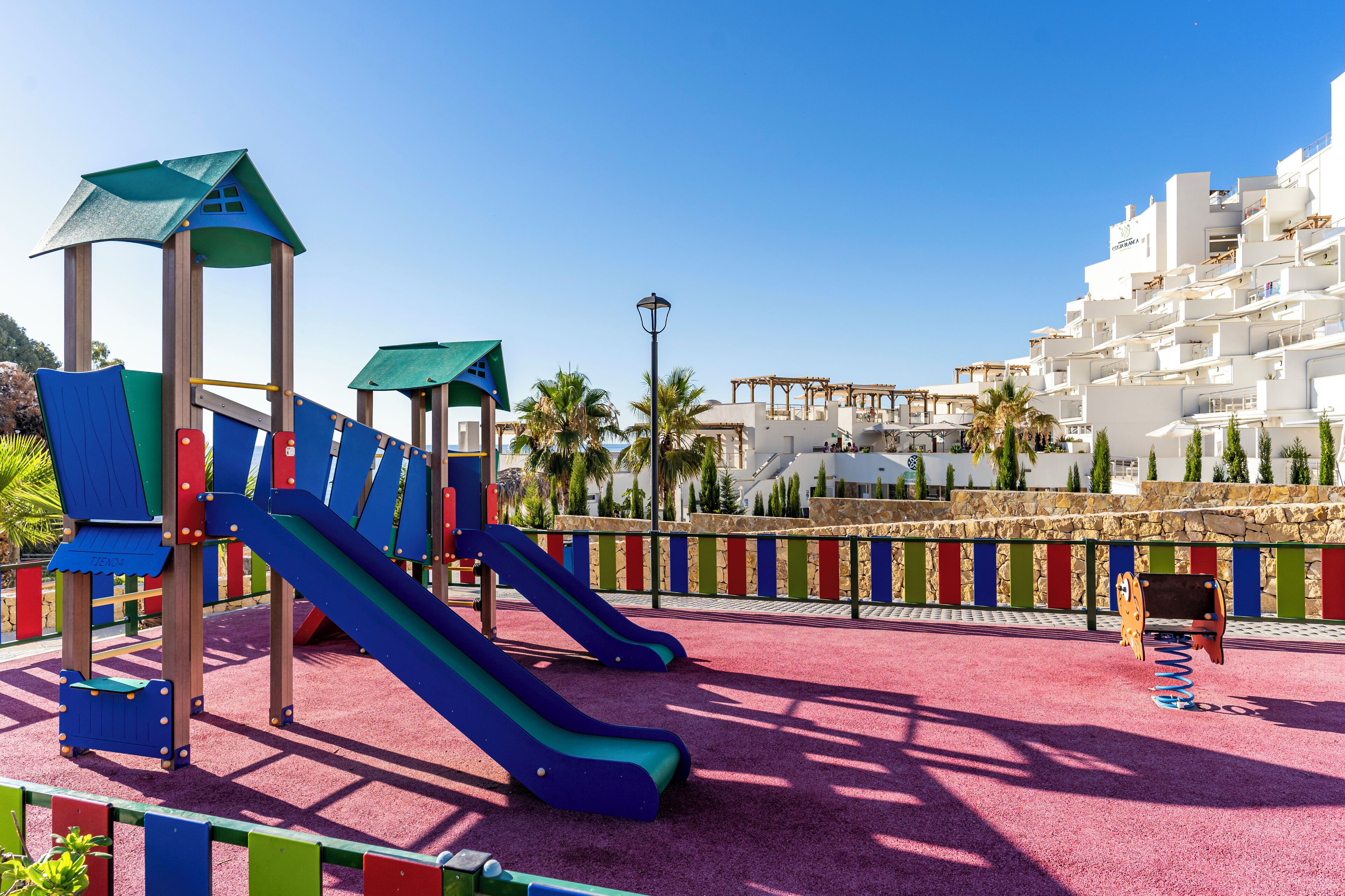 Un parque infantil con toboganes y estructuras para escalar, rodeado por una valla de colores y palmeras, con edificios blancos al fondo.