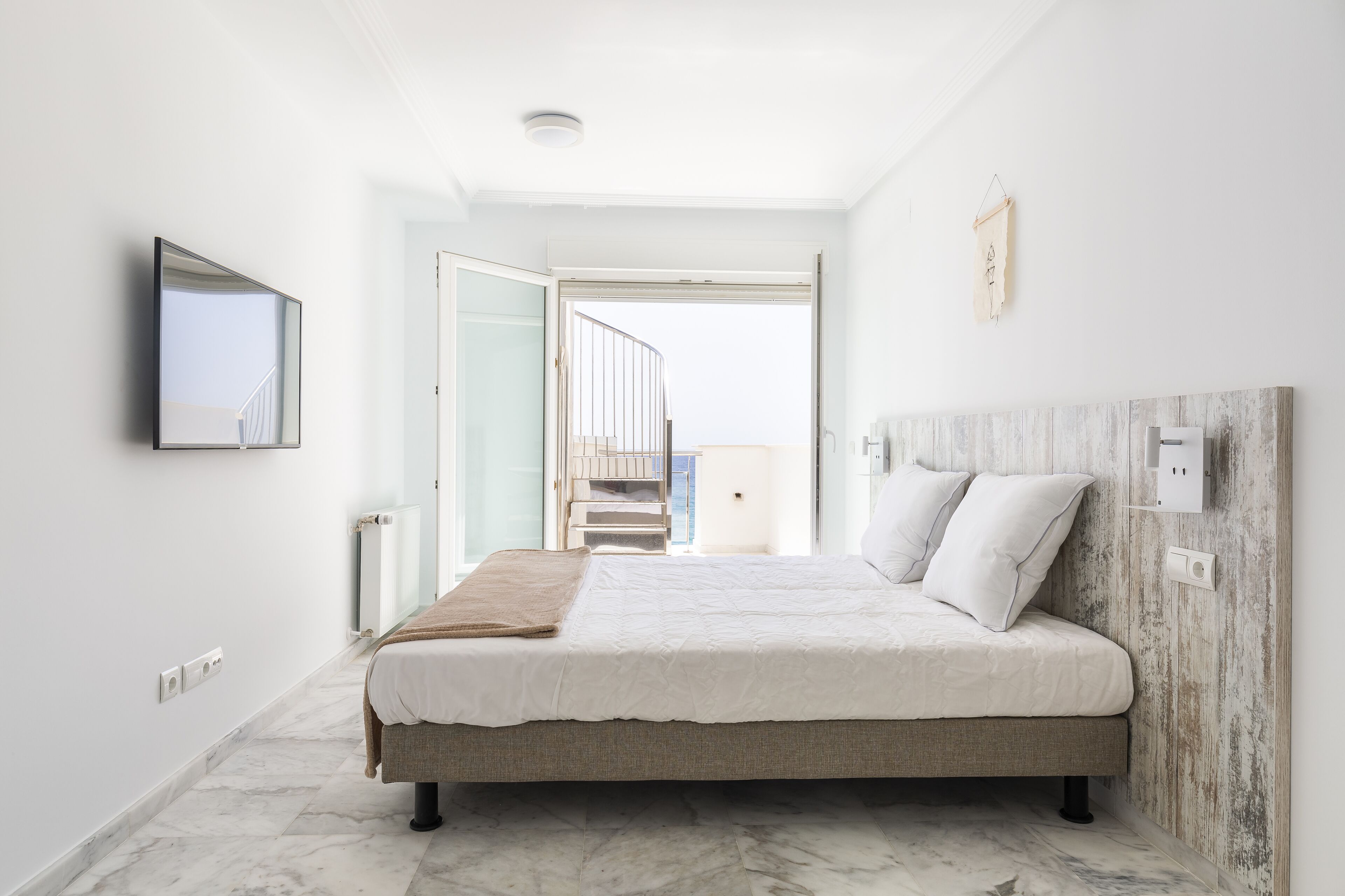 Een moderne slaapkamer met een groot bed, een houten hoofdbord, uitzicht op een balkon en een witte muur met een ingelijste foto.