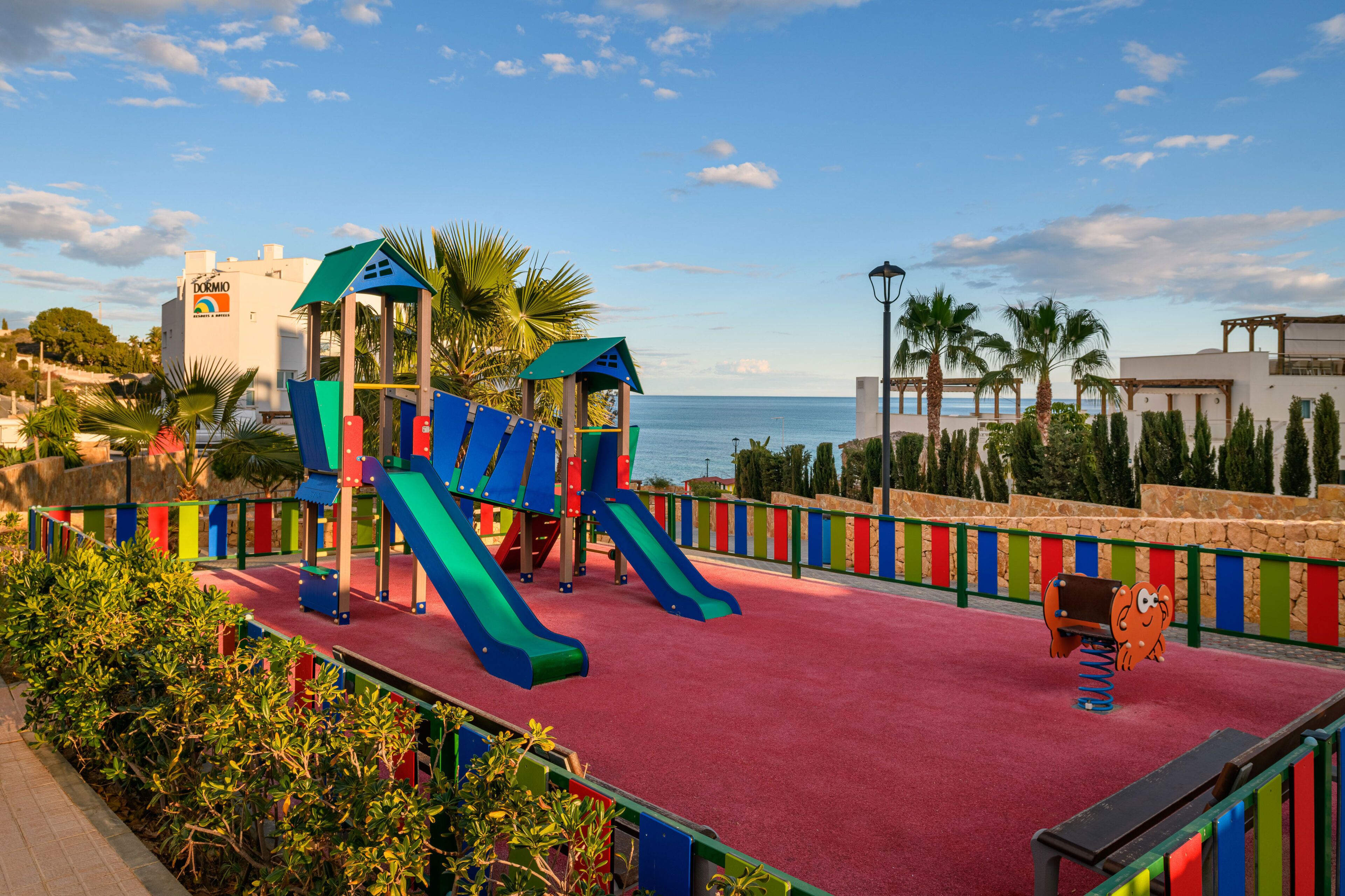 A playground with slides and climbing structures, a red carpeted area, and a view of the sea.