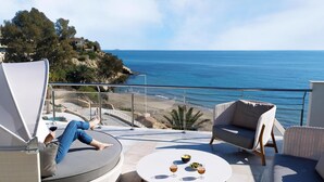 Een balkon met uitzicht op zee, een ronde tafel met glazen en een schaal, en een ligstoel.