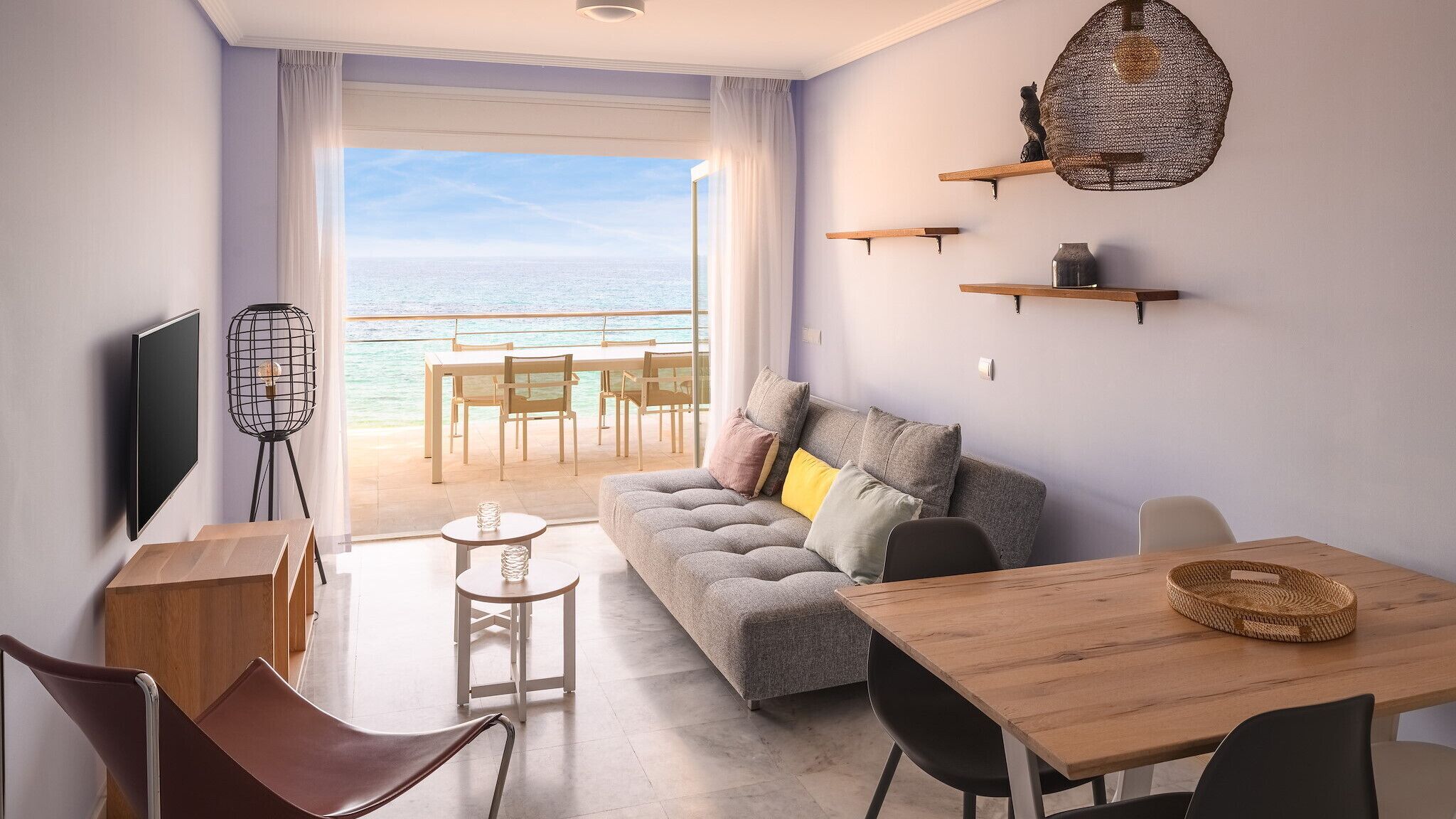 A modern living room with a sofa, a dining table, a TV, and a view of the sea.
