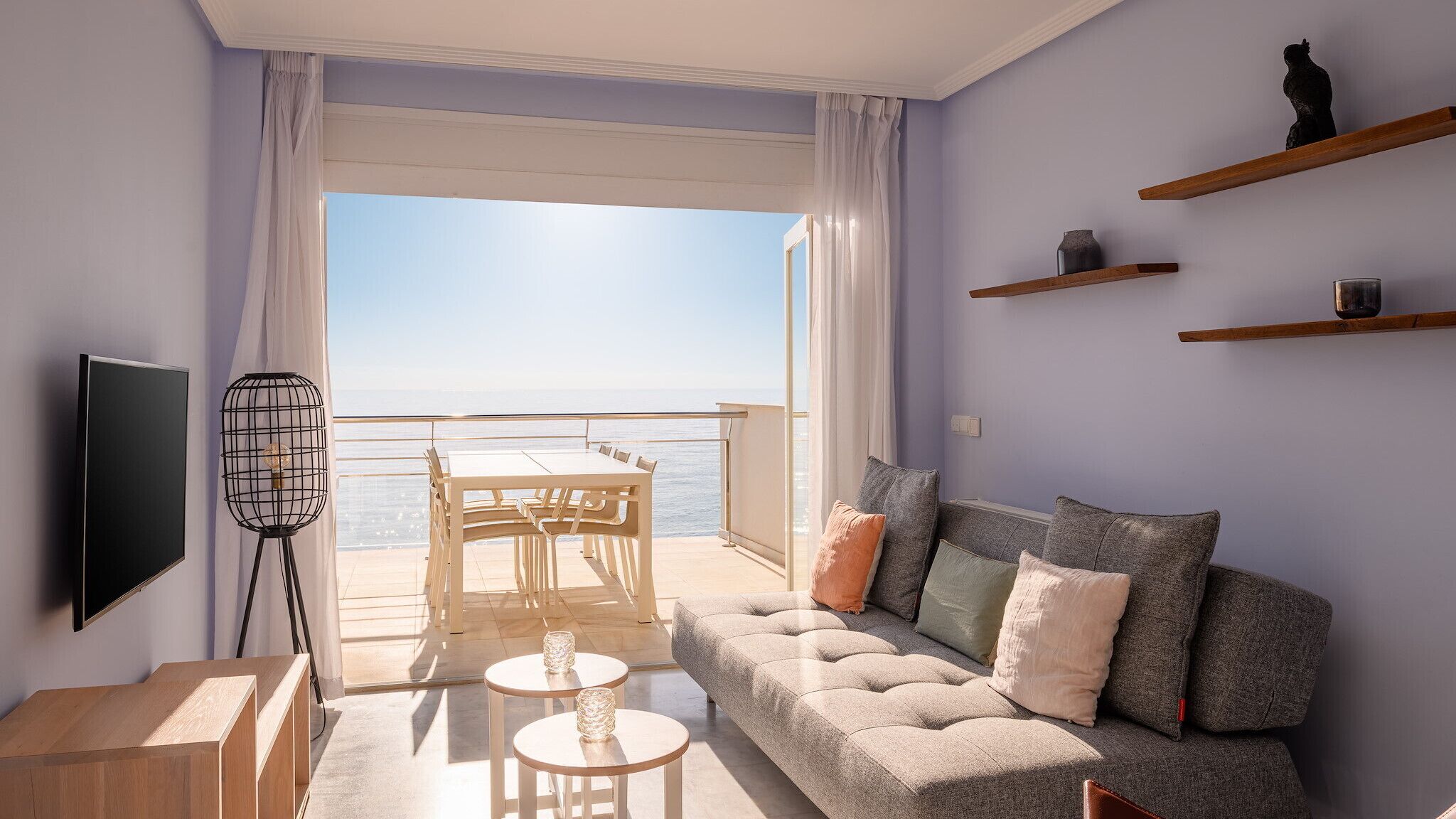 Una sala de estar moderna con balcón con vista al mar, un sofá con cojines, un televisor y una lámpara de pie.