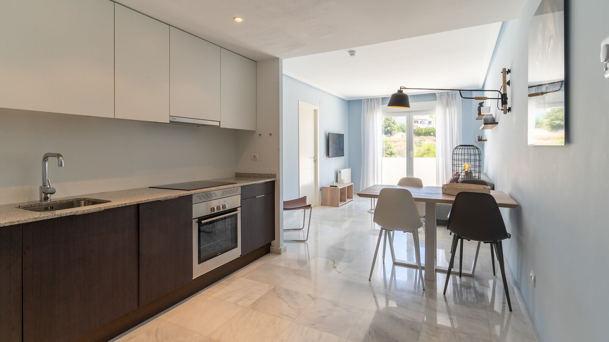 A modern kitchen with dark cabinetry, a stainless steel sink, and an oven. There is a dining area with a table and chairs.