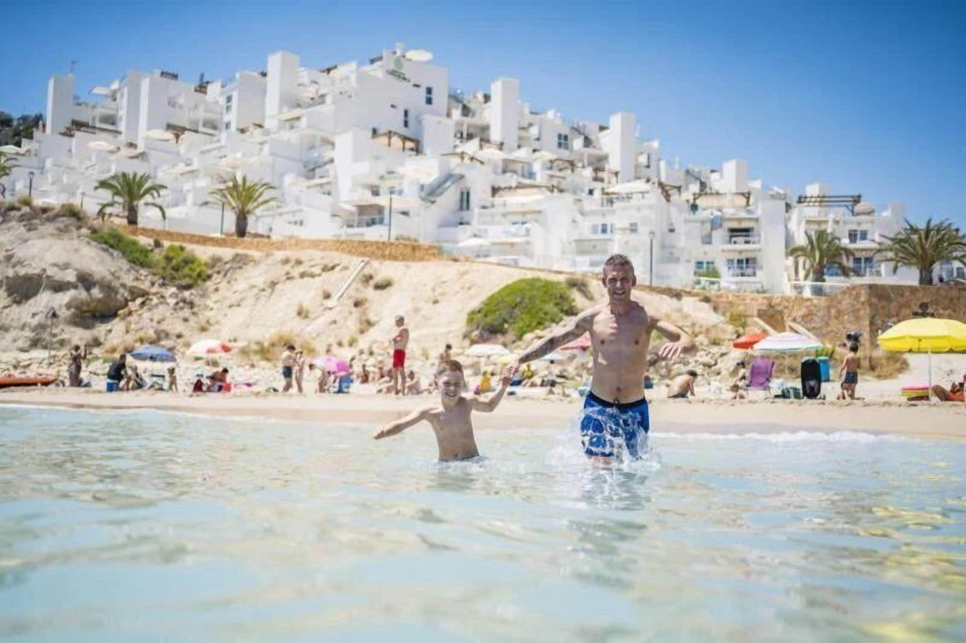 Una playa con edificios blancos al fondo, agua cristalina y gente disfrutando.