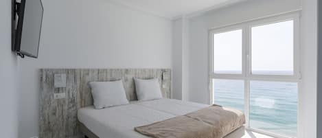 Quarto com cama grande, televisão fixada na parede e vista para o mar através de amplas janelas.