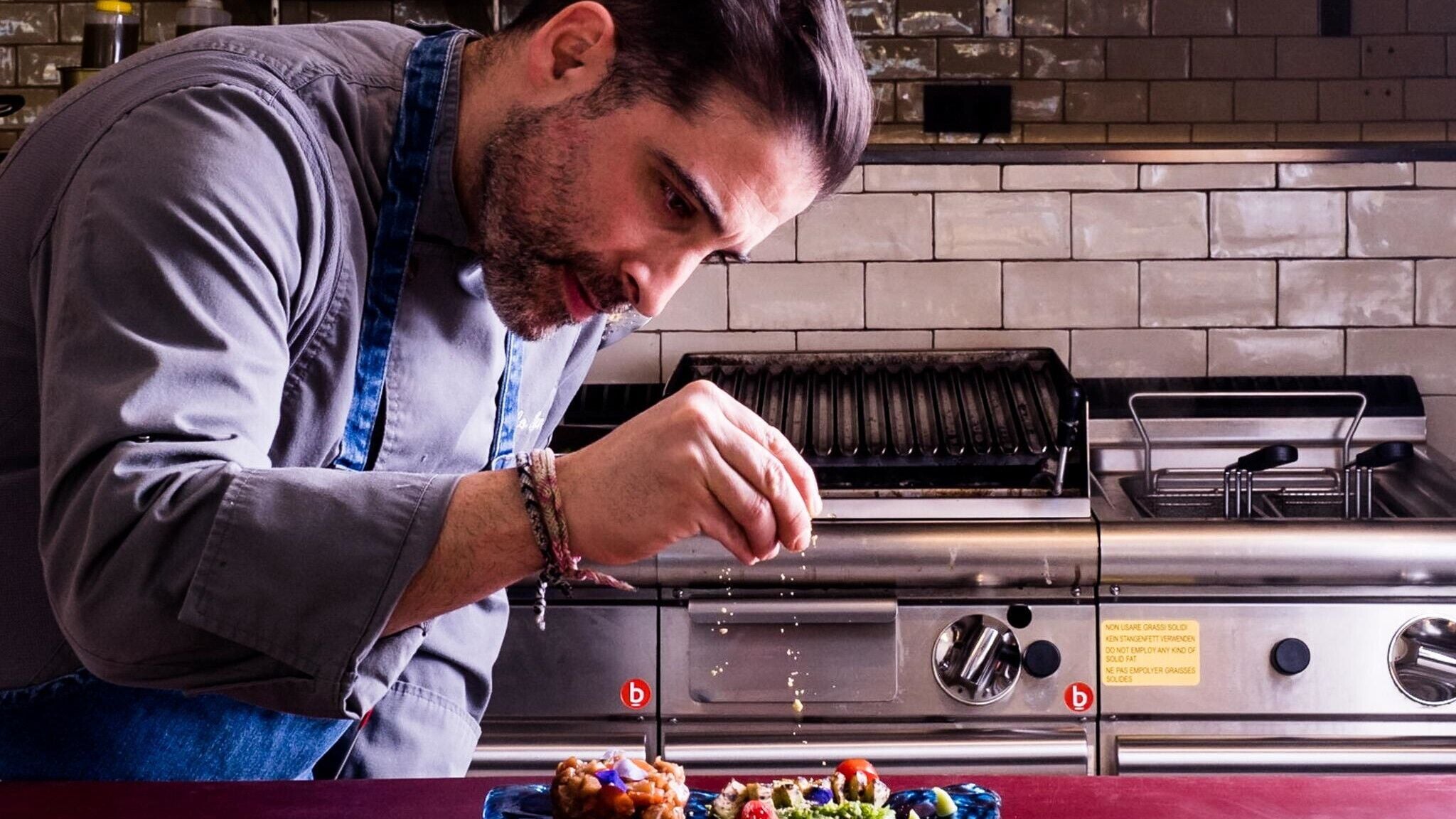 Un chef preparando comida en una cocina industrial con electrodomésticos de acero inoxidable y un salpicadero de azulejos.