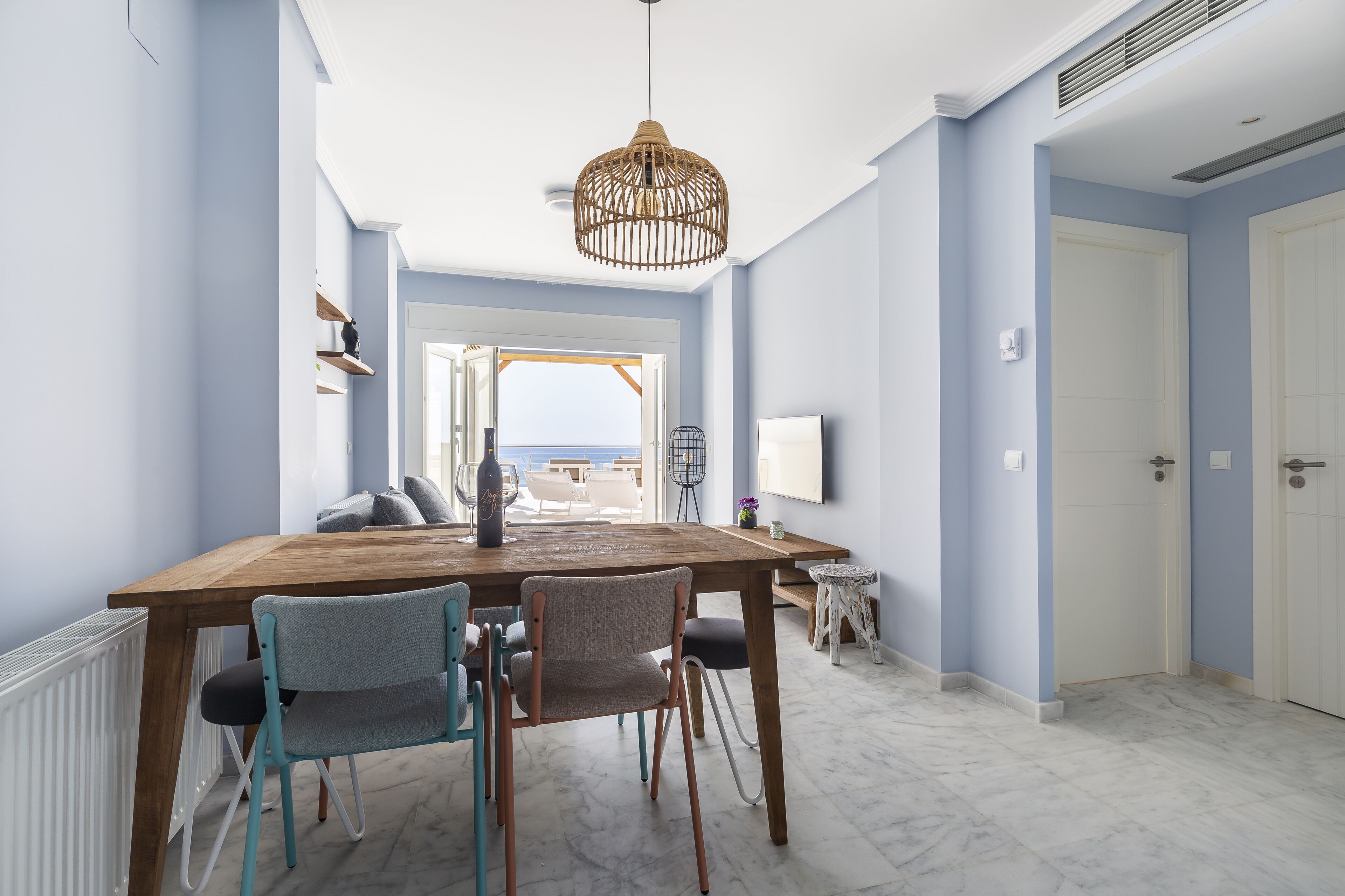 A dining area with a wooden table, two chairs, a hanging light fixture, and a view of the sea through an open door.