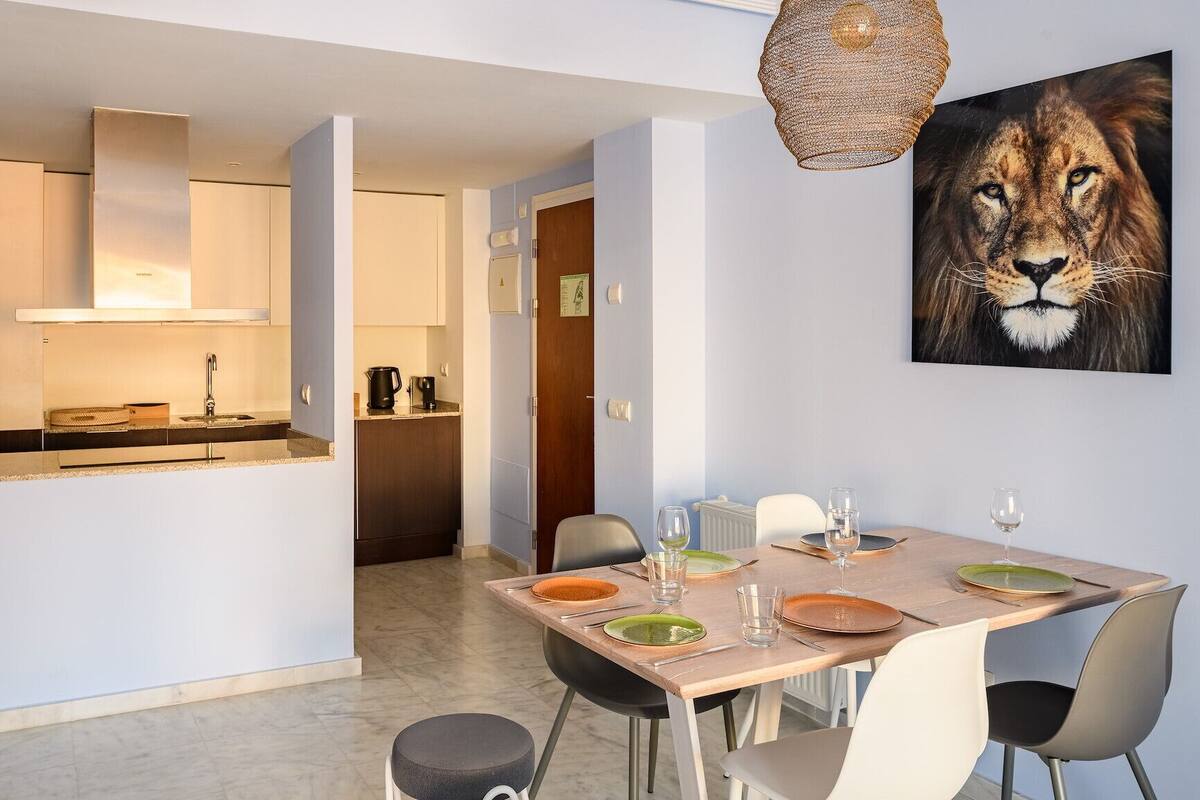 Een moderne keuken met een inbouwoven, een koffieapparaat en een spoelbak. Er is een eethoek met een tafel gedekt voor twee personen, stoelen en een schilderij van een leeuw aan de muur.