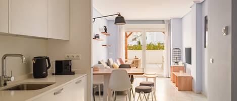 Une cuisine moderne équipée d’un four encastré, d’une machine à café et d’un évier. On y trouve également un coin repas avec une table et des chaises, ainsi qu’un balcon où l’on peut voir des plantes à travers les portes vitrées.